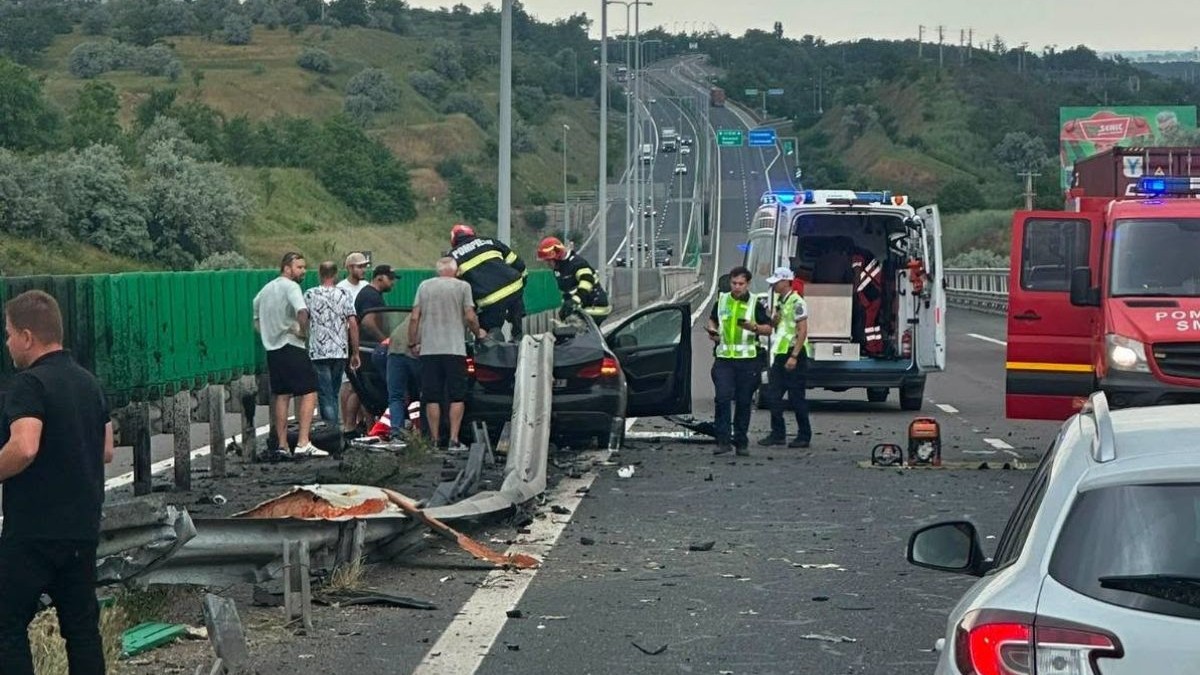 Imaginea surprinde scena unui accident rutier grav produs pe autostrada Soarelui A2, într-o zonă de pod sau viaduct flancată de dealuri cu vegetație. În centrul cadrului, un autoturism puternic avariat este oprit de-a curmezișul benzii, lipit de un parapet metalic separator care este complet deformat și rupt în urma impactului, iar pe asfalt sunt împrăștiate numeroase resturi. În jurul mașinii distruse s-au strâns mai mulți bărbați în haine civile, alături de care intervin direct pe vehicul doi pompieri echipați cu căști și costume de protecție. Pe banda din dreapta este oprită o ambulanță cu ușile din spate larg deschise, în apropierea căreia stau două persoane în uniforme și veste reflectorizante, iar pe jos, lângă salvare, a fost așezat un echipament tehnic portabil cu motor, probabil pentru descarcerare. În extremitatea dreaptă a imaginii se observă parțial o autospecială de pompieri de mari dimensiuni, cu portiera deschisă. În prim-plan, în colțul din dreapta jos, apare spatele unui alt autoturism oprit, pe marginea stângă a sensului de mers se află un panou înalt de protecție, iar șoseaua se continuă în fundal cu o porțiune lungă în rampă, mărginită de stâlpi de iluminat, pe care circulă alte vehicule aflate la distanță, 12 iunie 2026