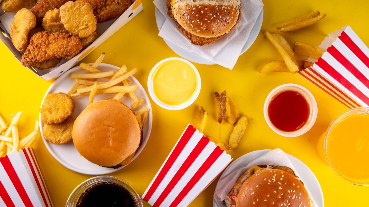 Imaginea arată o selecție variată de alimente tip fast-food, precum burgeri, cartofi prăjiți, nuggets și sosuri, dispuse pe o masă galbenă. Preparatele sunt bogate în grăsimi și ingrediente ultra-procesate, ilustrând perfect conceptul de mâncare rapidă și nesănătoasă.