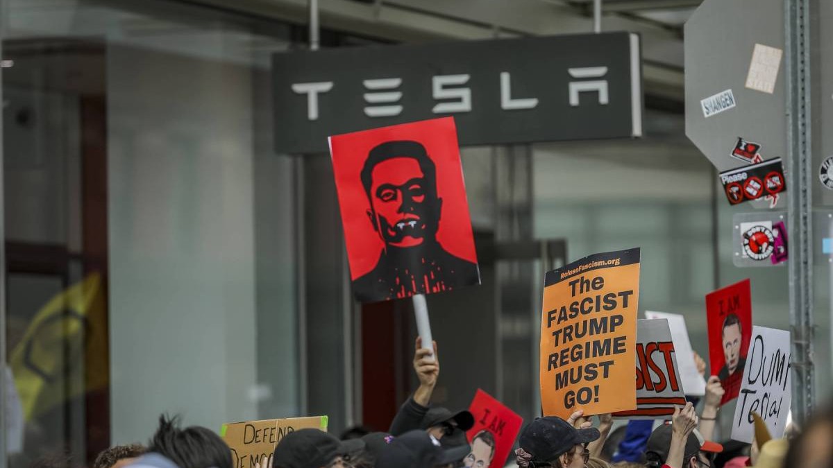 multime de oameni in fata unei reprezentante tesla cu pancarte cu mesaje impotriva lui elon musk si administratiei trump