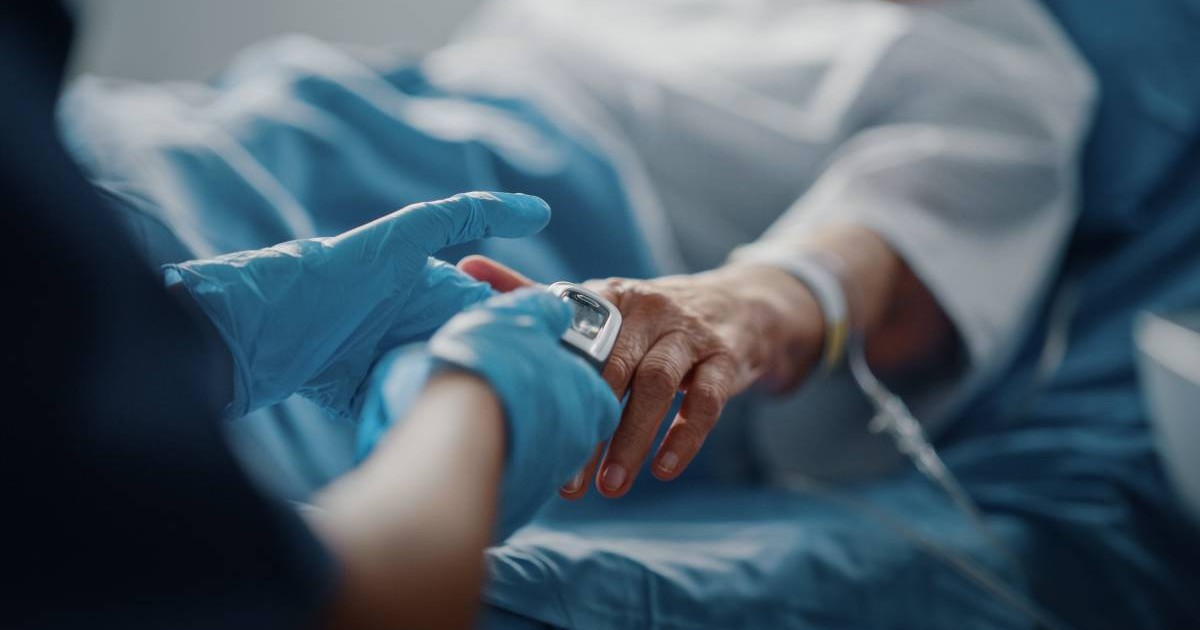 O femeie în vârstă pe patul de spital, după o intervenție, are puls oximetru montat la mână, iar o asistentă o îngrijește.