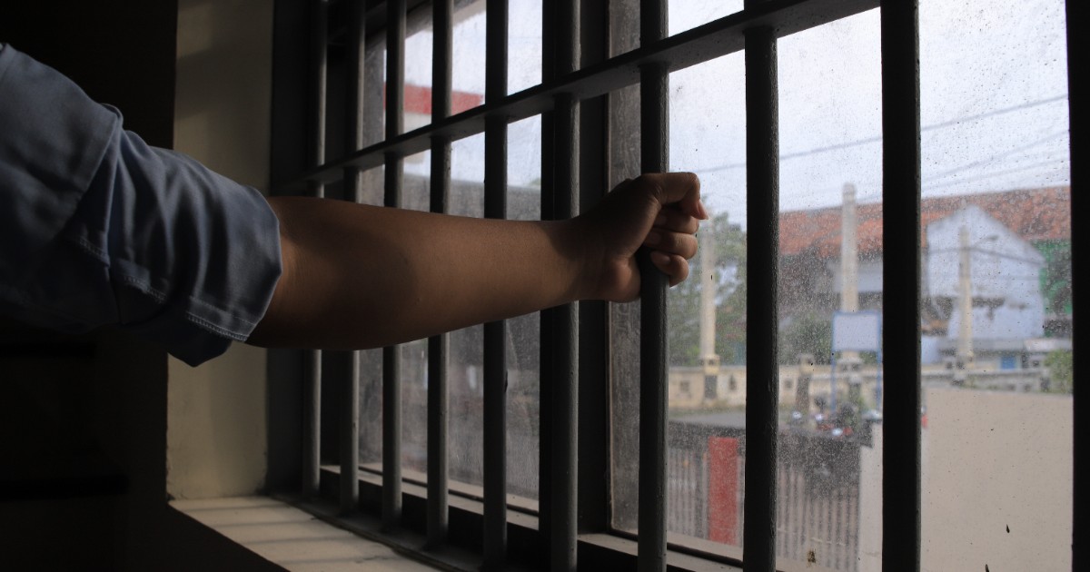 Un bărbat care stă în închisoare și se uită pe fereastră, afară este înnorat, iar el se ține cu mâinile de gratiile închisorii