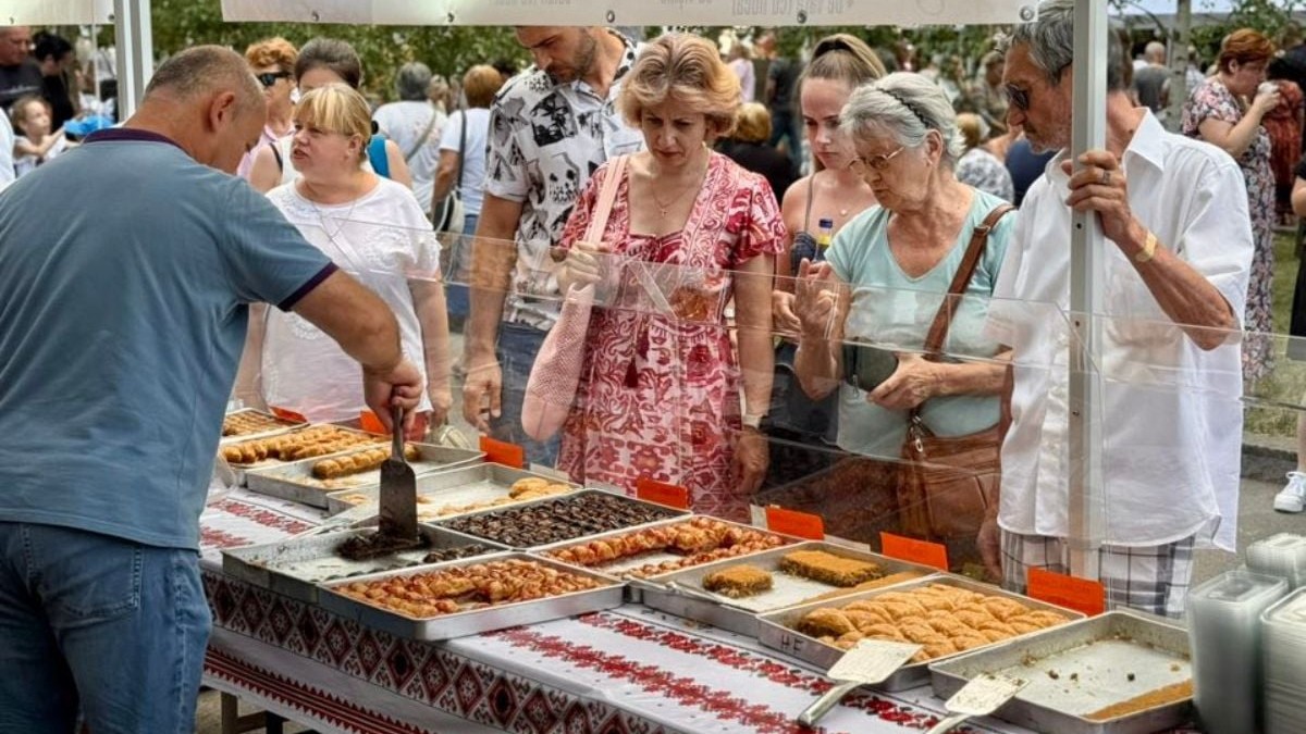 În imagine sunt vizibile mai multe tăvi metalice așezate pe o masă lungă, fiecare tavă conținând diferite tipuri de prăjituri sau produse de patiserie turcești. În partea stângă, un bărbat poartă un tricou bleu și ține o spatulă metalică deasupra unei tăvi. De cealaltă parte a mesei, câteva persoane stau grupate în așteptare, unele ținând genți sau sacoșe, spatele lor fiind acoperit parțial de o bară transparentă de protecție