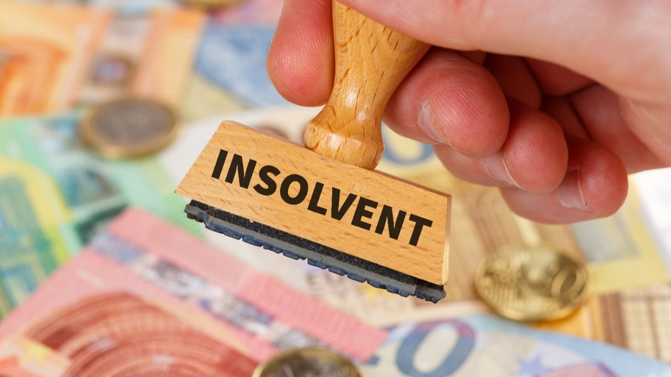 O mână care ține un ștampilă de lemn inscripționată cu cuvântul „INSOLVENT” deasupra unui fundal alcătuit din mai multe bancnote și monede euro.