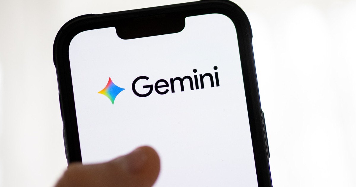 Imaginea este un prim-plan al ecranului unui smartphone cu ramă neagră, care afișează logo-ul și numele "Gemini" în litere negre, pe un fundal alb strălucitor. Ecranul este ținut de o persoană a cărei mână și deget sunt vizibile în partea de jos a cadrului.