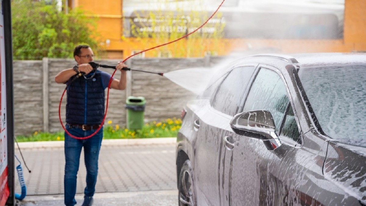 Greșeala pe care o fac șoferii când spală mașina la spălătoriile Self-Wash - Un bărbat clătește un autoturism  gri cu jet de apă sub presiune la o spălătorie 