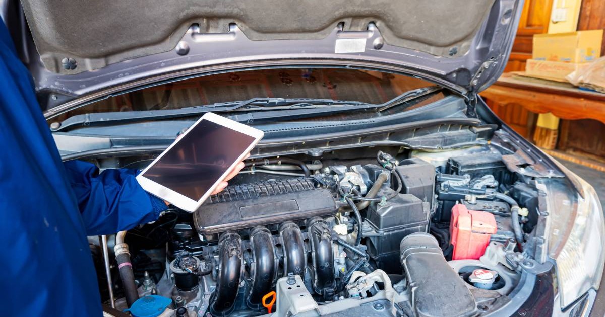 mecanic auto care tine in mana o tableta alba, in fata unei masini care are capota deschisa, posibil intr-un service auto