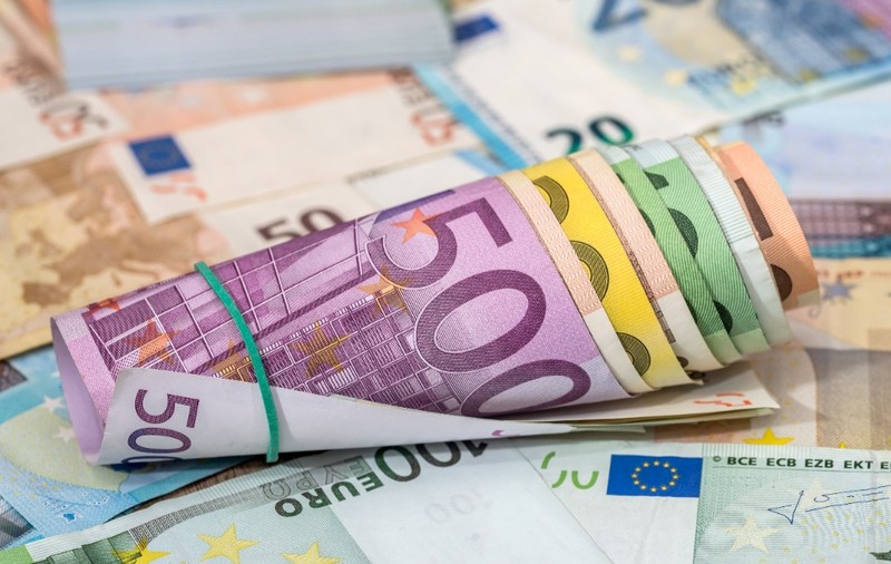 Un sul de bancnote euro de diferite cupiuri, prins cu un elastic verde, așezat peste alte bancnote euro împrăștiate.