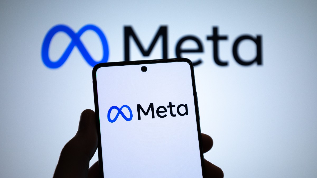 Imaginea arată un smartphone ținut în mână, pe ecranul căruia este afișat logo-ul Meta, având în fundal același logo mărit și ușor estompat. Compoziția sugerează focusul companiei pe tehnologie mobilă și pe prezența sa dominantă în ecosistemul digital.
