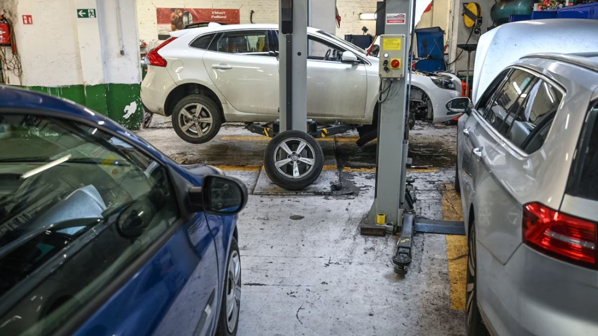 O mașină albă este în service auto, fără o roată, în fundal, iar în lateral se văd alte două mașini, una albastră și una gri