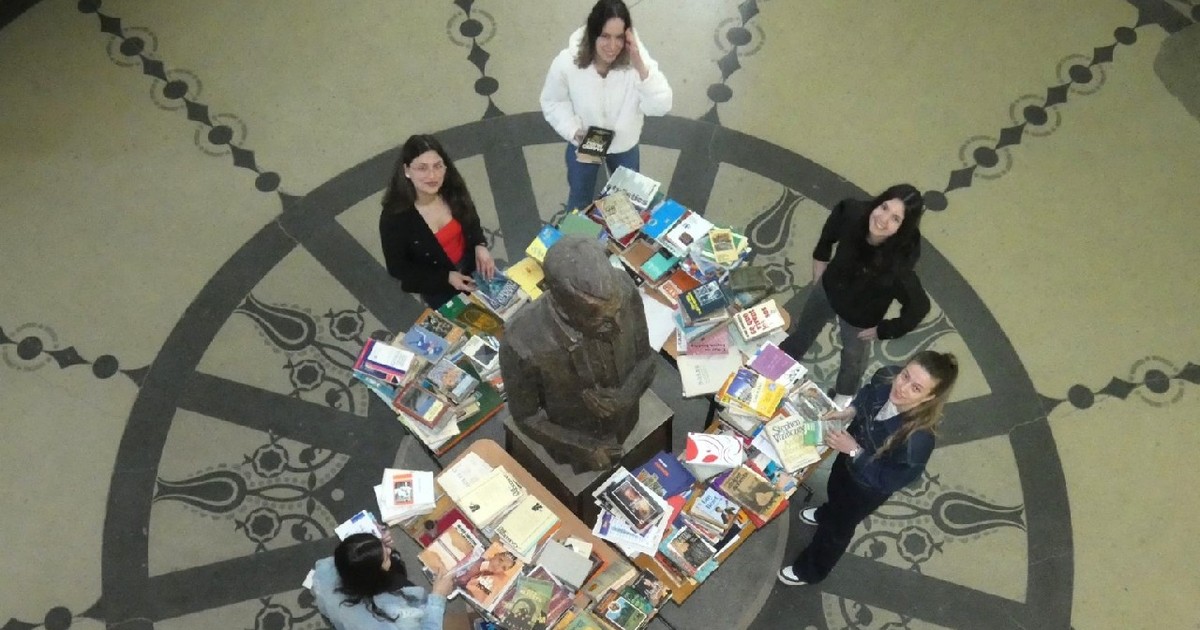 Mai multe studente se uita la o masă cu cărți, pusă în mijlocul unui hol