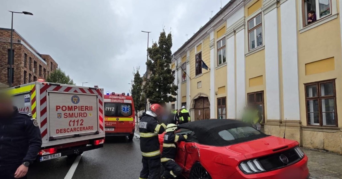 O mașină sport roșie, decapotabilă, prezintă avarii la portiera șoferului. La fața locului sunt pompieri SMURD, cu cel puțin două autospeciale vizibile.