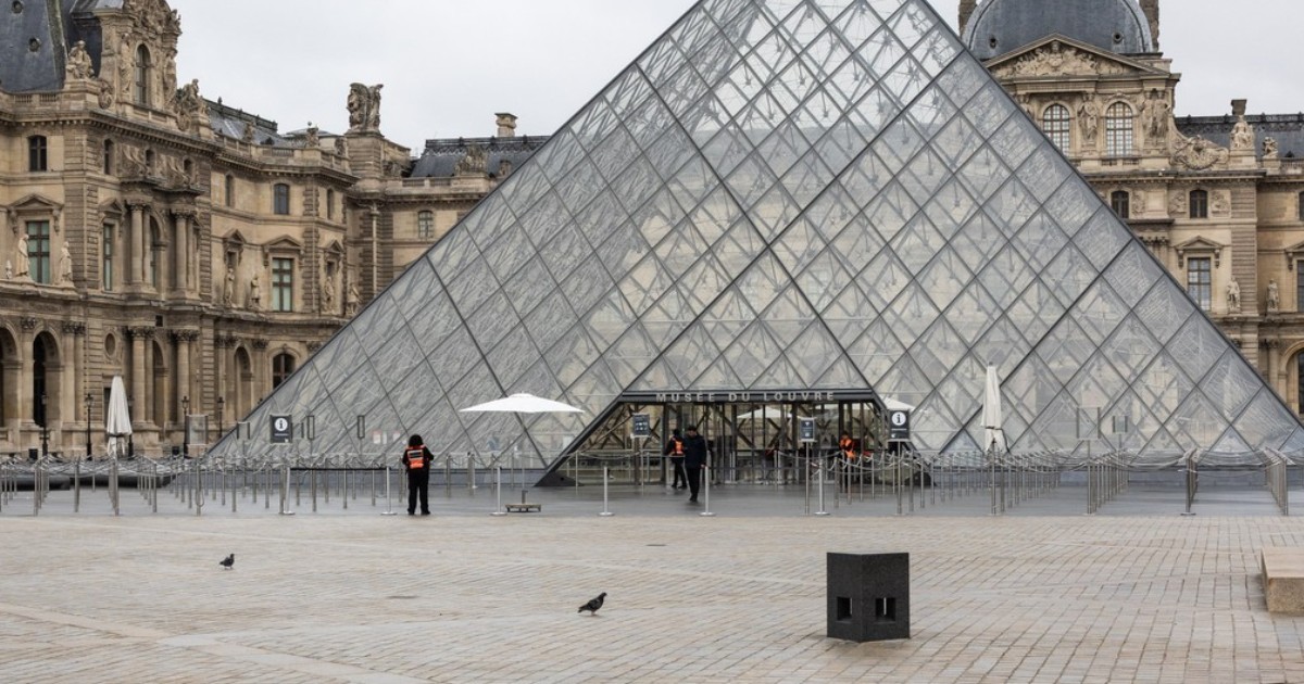 Intrare principală la Muzeul Luvru din Paris, înconjurată de clădirile istorice ale muzeului în fundal. Pe fundalul cerului înnorat se observă câteva persoane la intrare și câteva păsări pe pavaj
