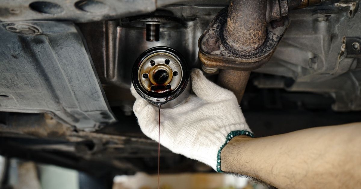Mâna unui mecanic auto ține un filtru de ulei, sub motorul unei mașini, din care se scurge ulei negru de motor
