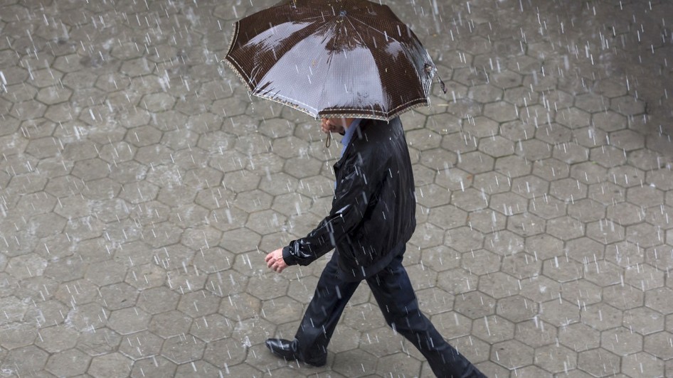 Un bărbat îmbrăcat într-o geacă neagră și pantaloni închiși la culoare, mergând pe un trotuar pavat în timp ce ține o umbrelă maro deasupra capului, pe o ploaie torențială.
