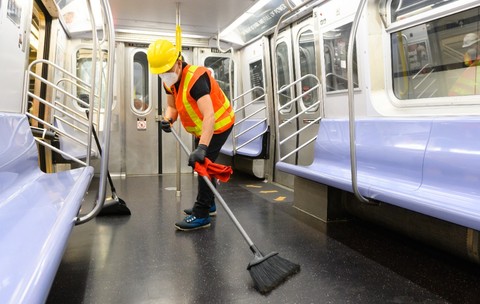 Un angajat la curățenie purtând cască de protecție, vestă reflectorizantă și mască mătură podeaua în interiorul unui vagon de metrou.   
