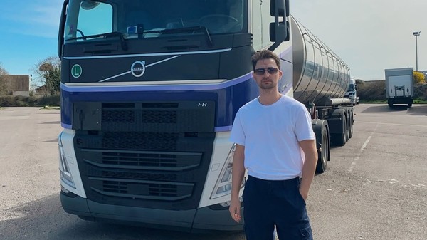 Un bărbat îmbrăcat în tricou alb, cu ochelari de soare, în fața unui camion cisternă Volvo într-o parcare.