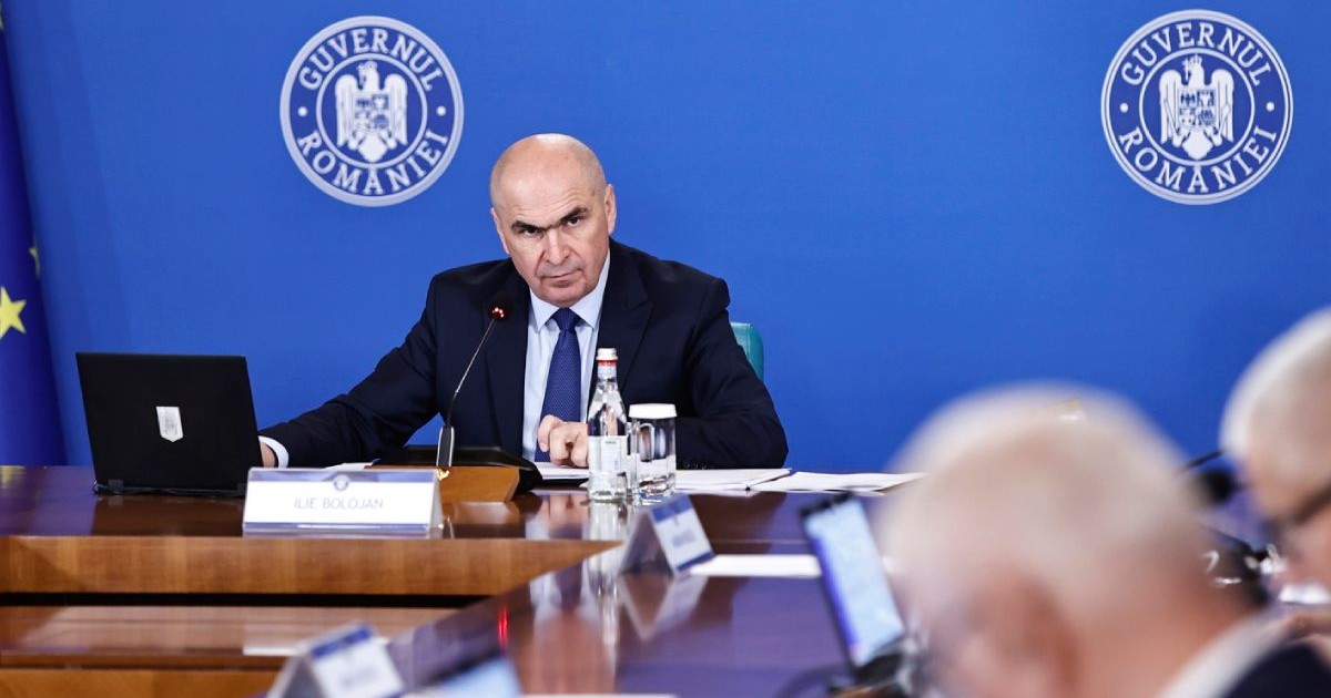 Ilie Bolojan, premierul României, îmbrăcat în costum negru, cu cămașă albă și cravată albastră, pe scaun, la ședința de guvern