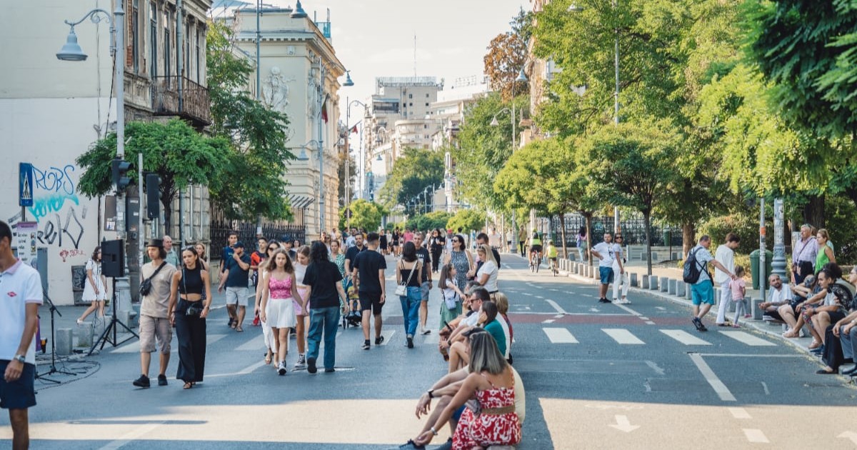 Calea Victoriei din București, într-o zi însorită. Oamenii au ieșit la plimbare, drumul este pietonal, iar unele persoane s-au așezat și stau de vorbă pe bordură