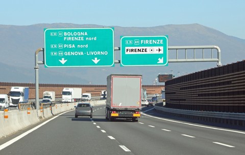 Traficul de pe o autostradă din Italia, cu mai multe autovehicule și camioane deplasându-se pe benzi sub panouri verzi mari de semnalizare rutieră ce indică direcțiile spre Bologna, Firenze, Pisa, Genova și Livorno.