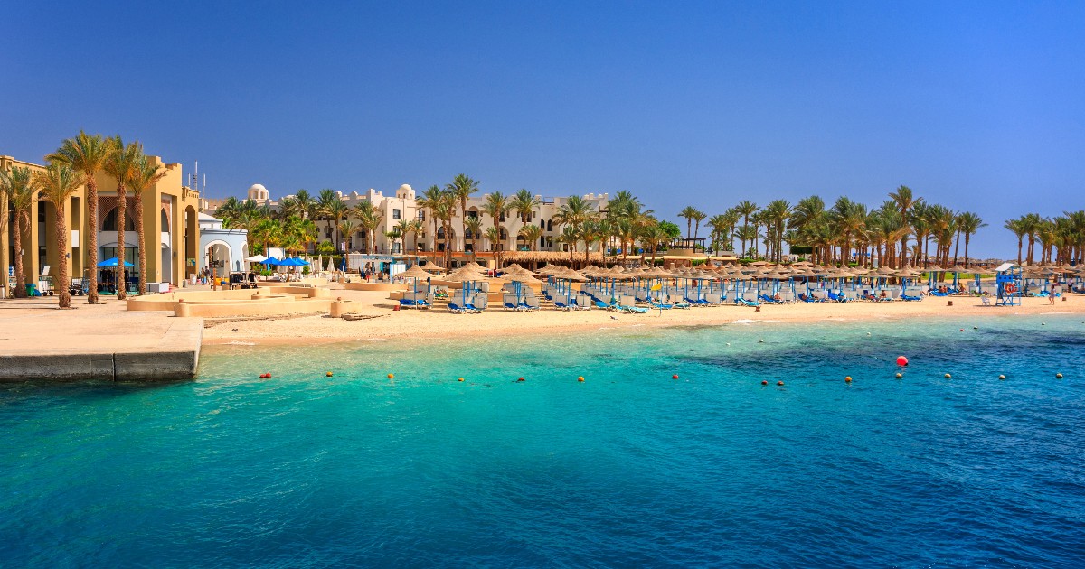O plajă dintr-un resort din Egipt, cu nisip alb, mărginita de palmieri și de o clădire mare, în stil mediteranean. Marea, de un albastru intens, are ape cristaline în apropierea țărmului, în timp ce în larg se observă o nuanță mai închisă, de un albastru marin profund.
