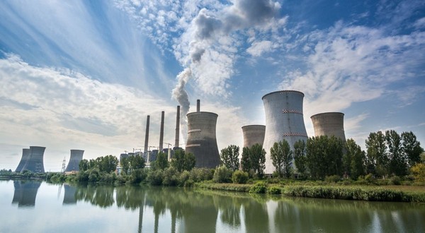 Termocentrala Turceni, cu un lac în față, turnurile uriașe pe fundal pe care iese fum și cerul albastru cu nori albi