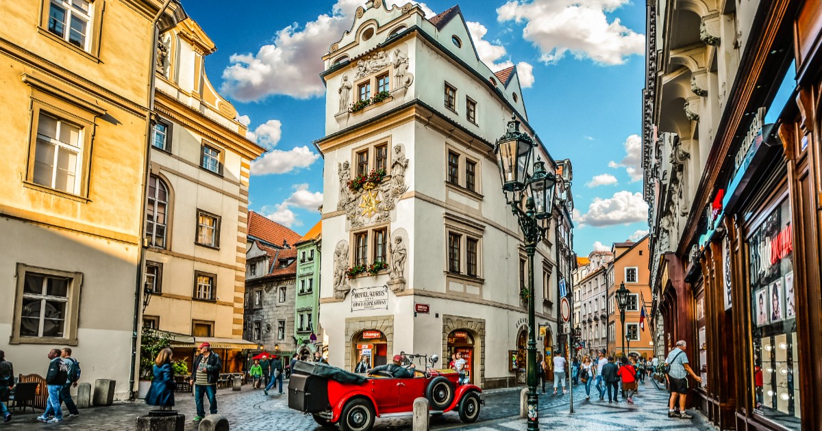 O stradă din centrul orașului Praga, mașinile sunt parcate pe marginea drumului, o mașină retro, roșie, este parcat chiar în fața unei intrări într-un imobil istoric