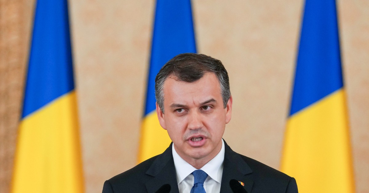Imaginea îl surprinde pe Eugen Tomac în timpul unei declarații oficiale, având în fundal mai multe drapele ale României. Politicianul poartă un costum închis la culoare și o cravată albastră, fiind fotografiat în timp ce se adresează publicului de la un pupitru cu microfoane.