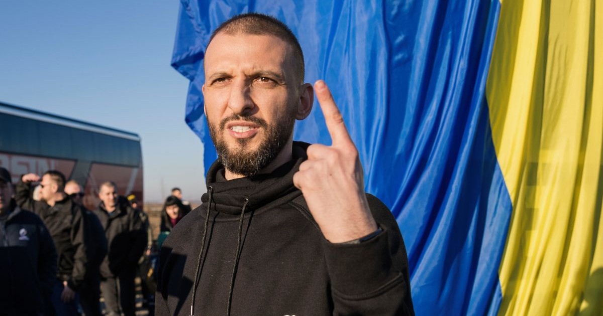 Stefan Mandachi cu barbă, intr-un hanorac negru cu scris pe el, tine degetul aratator de la mana stanga in sus, in spate un steag din care se vad albastru si galben si o multime de oameni, 24 ianuarie 2020