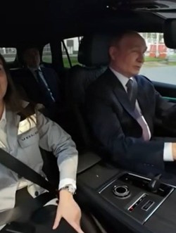 Vladimir Putin conduce un SUV Volga, îmbrăcat într-un costum închis la culoare și cravată. Pe scaunul din dreapta se află o femeie purtând centura de siguranță, iar în spate se vede un alt pasager. Imaginea este surprinsă din interiorul mașinii, în timp ce vehiculul se deplasează printr-o zonă industrială.