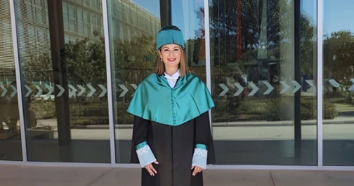 Imaginea prezintă o femeie zâmbitoare, îmbrăcată în haine academice de ceremonie (robă), stând în fața unei clădiri moderne cu pereți de sticlă.