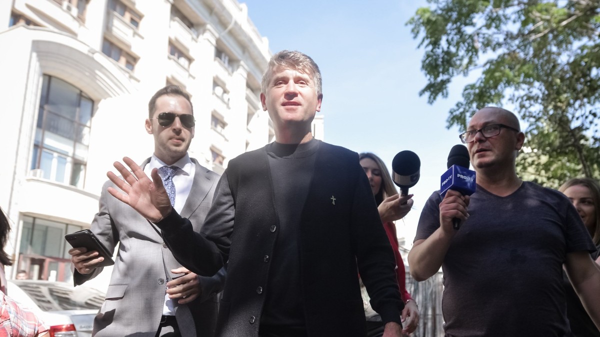 În imagine apare o persoană care se deplasează pe o stradă din oraș, înconjurată de mai multe persoane și jurnaliști care îi adresează întrebări. Fotografia surprinde un moment de interacțiune cu presa în spațiul public, pe fundalul unor clădiri și al unei zile însorite.