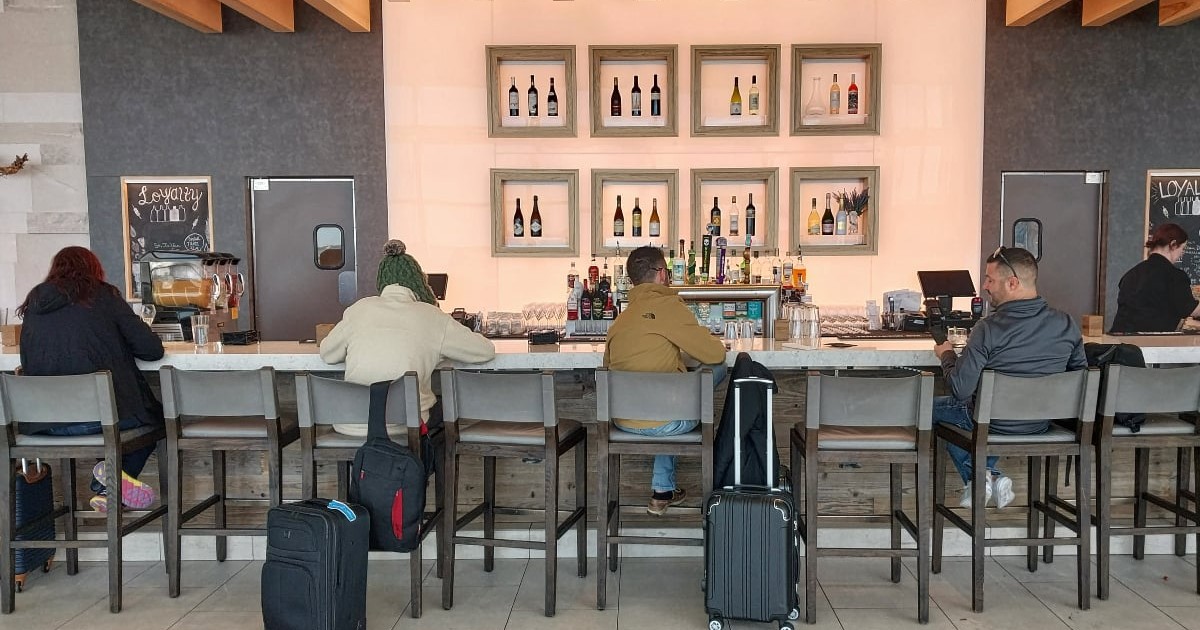 Mai mulți bărbați care stau pe scaune la un bar dintr-un aeroport, cu valizele lângă ei