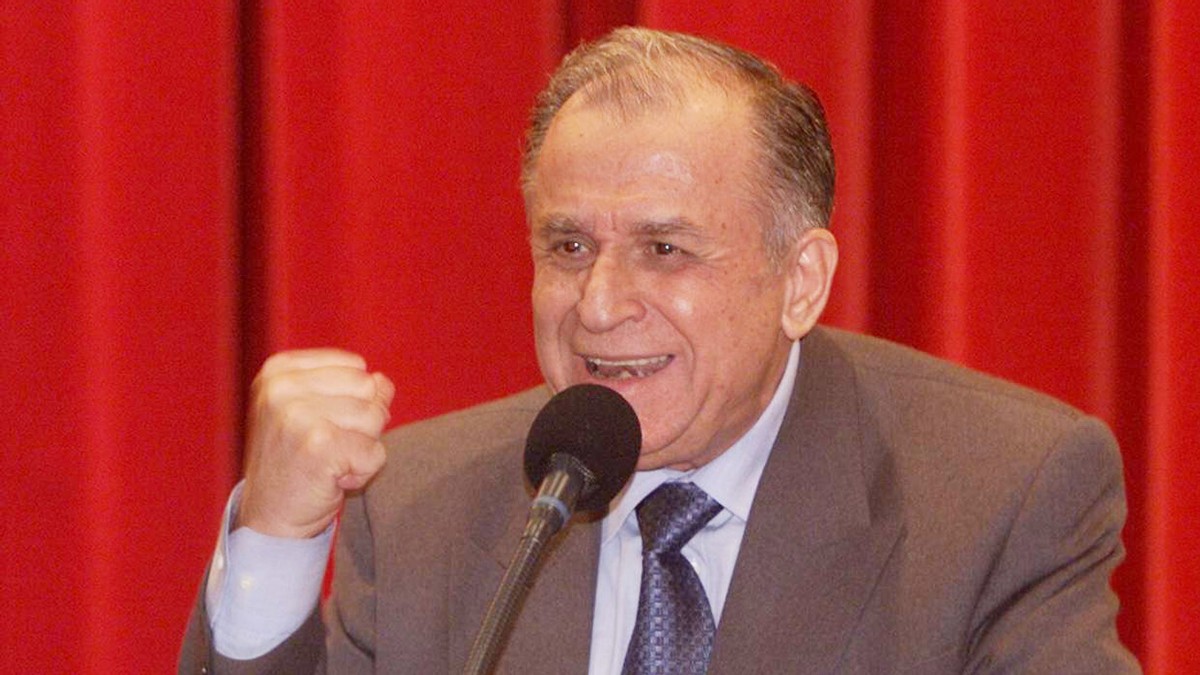 Ion Iliescu, fost președinte al României, îmbrăcat în costum gri, cravată albastră și cămașă albă. În spatele său, un fundal roșu