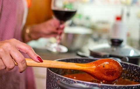 O femeie în bucătărie care gătește. În mâna stângă ține un pahar de vin