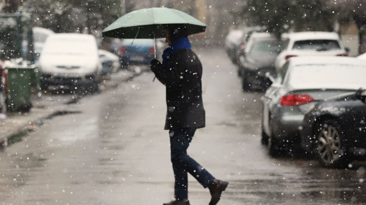 Un bărbat gros îmbrăcat care se acoperă cu o umbrelă traversează o stradă într-o zi în care ninge, București, 8 ianuarie 2025
