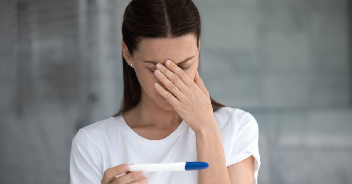 O femeie cu un test de sarcină în mână, și-a dus o mână la cap și privește tristă spre rezultatul indicat