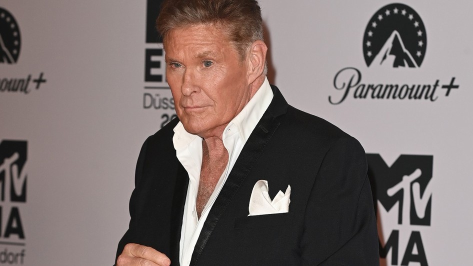 David Hasselhoff în costum negru și cămașă albă în timp ce face poză la un panou