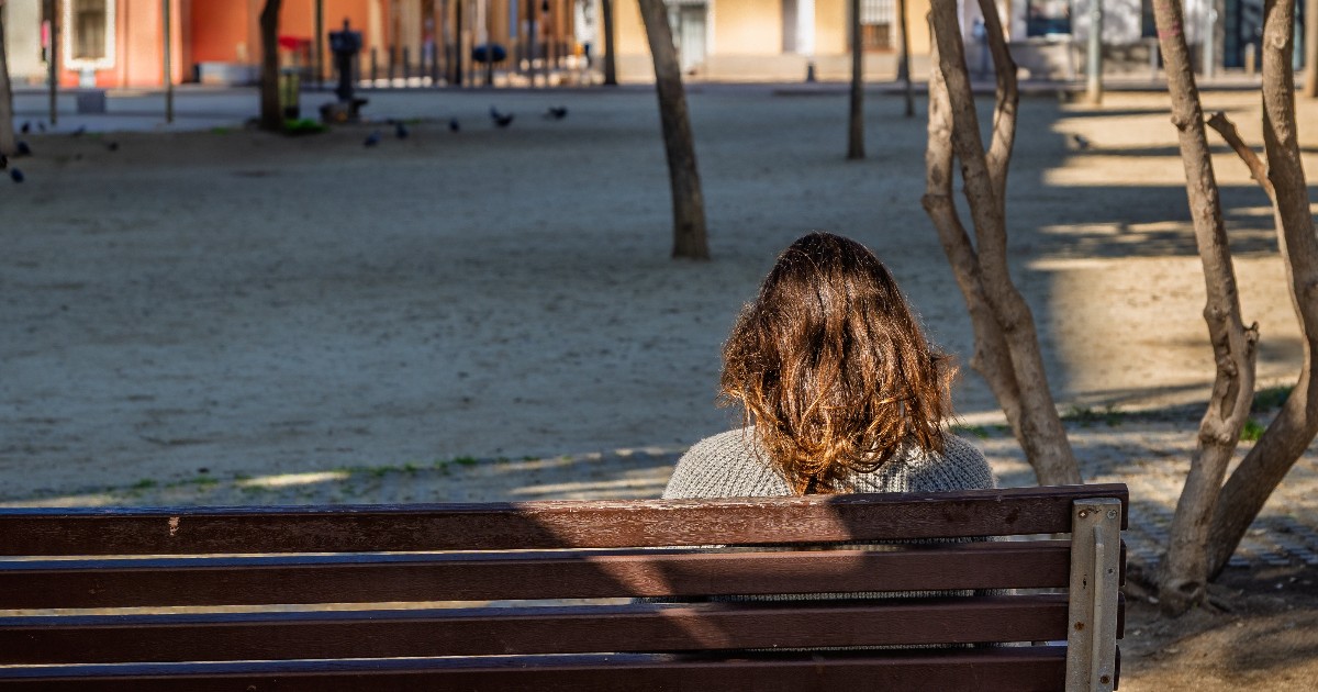 O tânără stă singură pe o bancă într-un parc, surprinsă din spate, într-un cadru liniștit, dar rece. Imaginea transmite izolare și reflecție, sugerând o situație dificilă sau un moment de cumpănă personală.