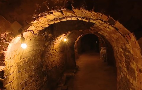 Interiorul unui tunel sau al unui beci subteran vechi, zidit din cărămidă roșie cu tavane boltite, iluminat de becuri.