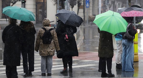 Mai mulți pietoni așteptând la o trecere de pietoni pe o vreme ploioasă și rece, adăpostindu-se sub umbrele colorate și purtând haine groase.