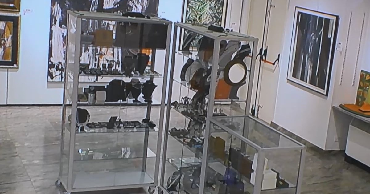 Interiorul unei galerii de artă sau case de licitații, unde se observă vitrine metalice cu geamuri transparente pline cu diverse obiecte decorative și bijuterii, în timp ce pereții din fundal sunt decorați cu numeroase tablouri și picturi abstracte.