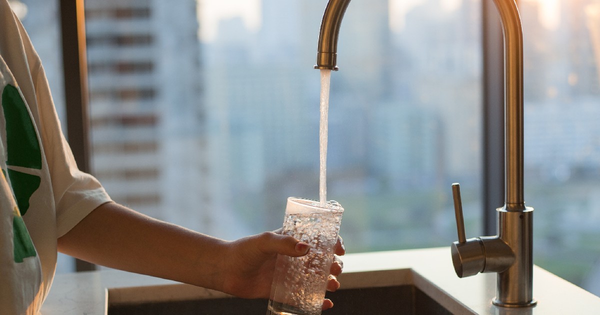 Fata umple un pahar cu apă de la robinet filtrată curată pentru băut în bucătărie acasă. Fereastră în fundal. Robinet modern și chiuvetă în bucătăria de acasă.