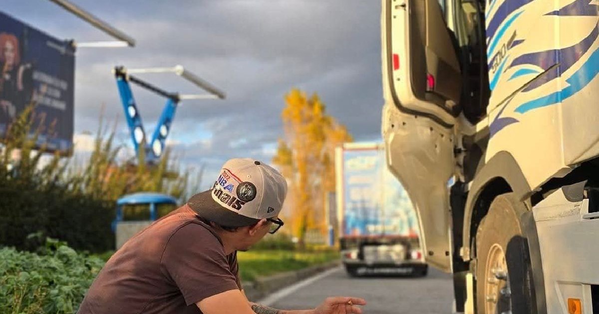 Un șofer de TIR cu șapcă pe cap stă pe o bordură, lângă camionul care are portiera deschisă