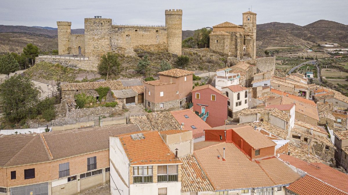 Fotografie a unui oraș istoric cu ruinele unui castel de piatră situat pe vârful unui deal, înconjurat de acoperișuri cu țigle roșii și clădiri cu pereți albi.