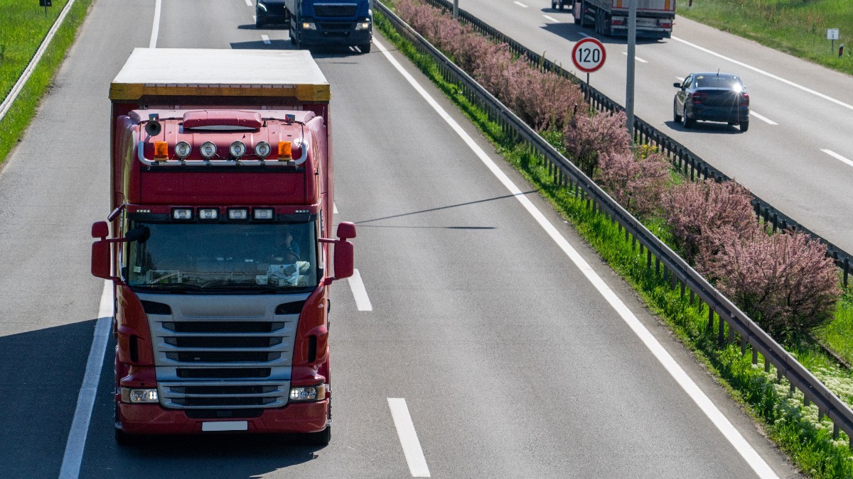 Un camion roșu rulând pe prima bandă a unei autostrăzi aglomerate, încadrat de alte autovehicule și un parapet median cu vegetație.