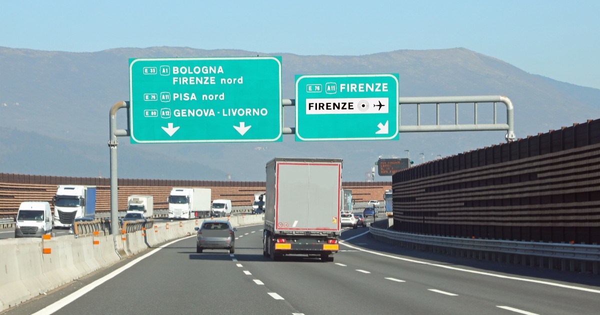 Imaginea prezintă traficul de pe o autostradă din Italia, cu vehicule aflate în mers sub două panouri verzi mari de semnalizare care indică direcțiile către orașe precum Bologna, Pisa și Firenze.