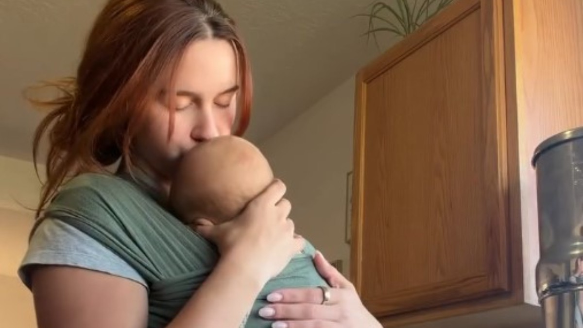 O tânără care sărută blând pe cap nou-născutul purtat într-un sistem de wrap textil verde, într-un cadru casnic.