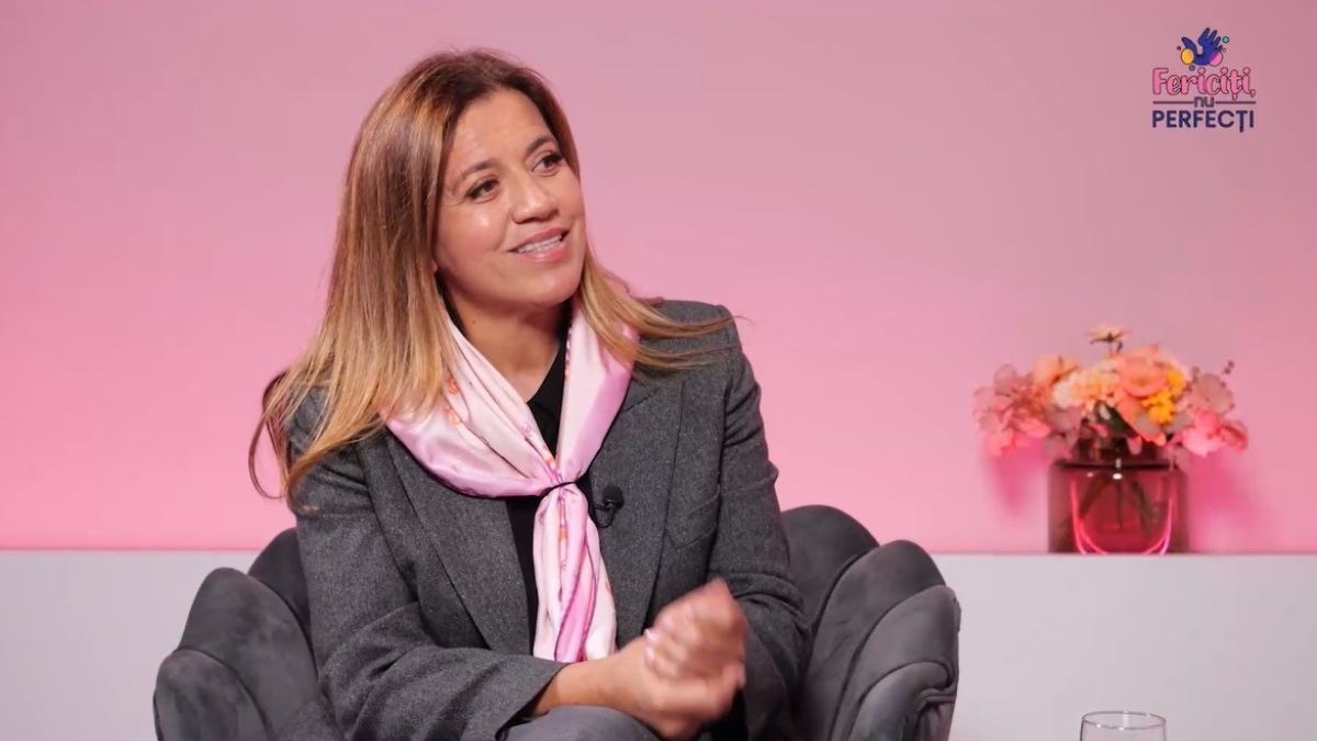 Mirela Retegan, îmbrăcată într-un costum gri, cu o eșarfă roz la gât, pe un fotoliu gri, iar pe fundal un perete roz și o vază cu flori