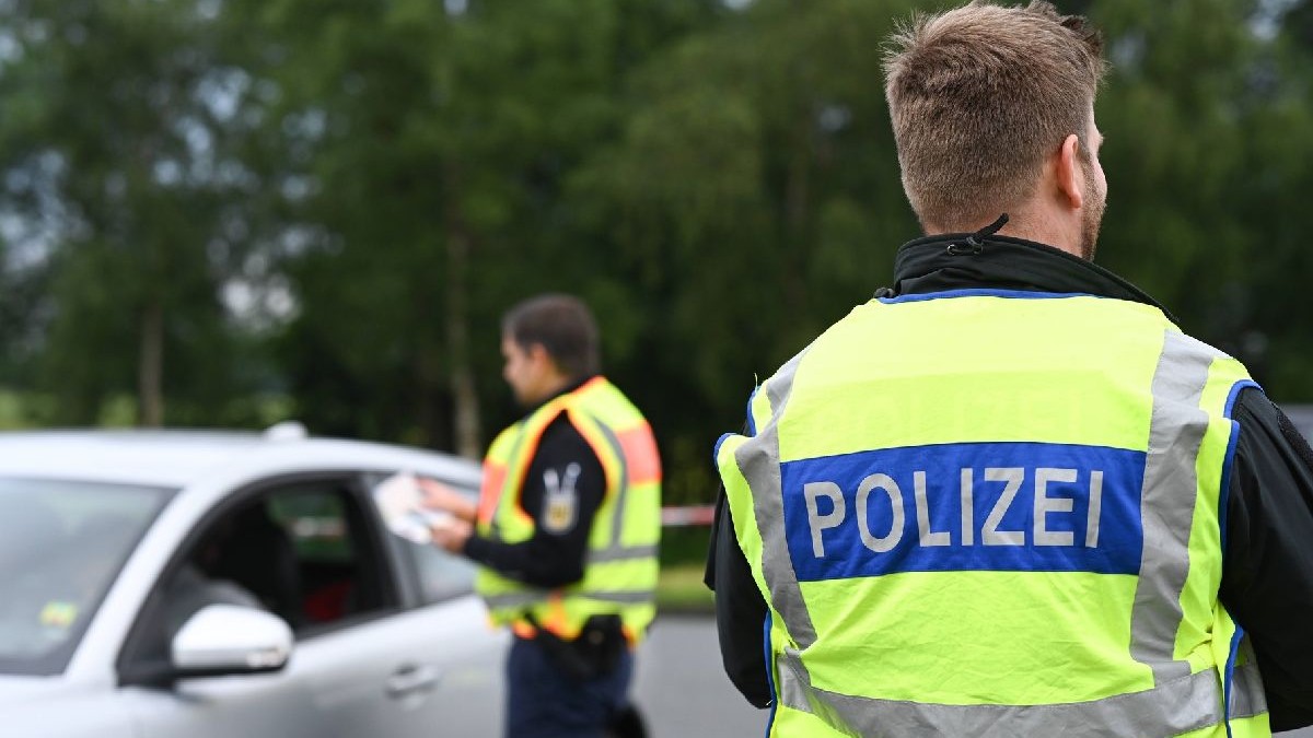 Fotografia surprinde doi polițiști în timpul unui control rutier. Polițistul din prim-plan poartă o vestă galben cu albastru cu inscripția „POLIZEI” în litere albe îngroșate, este văzut din spate, în fața unei mașini argintii. Al doilea polițist, din fundal, conversează cu o persoană din interiorul mașinii. În fundal se văd copaci verzi.