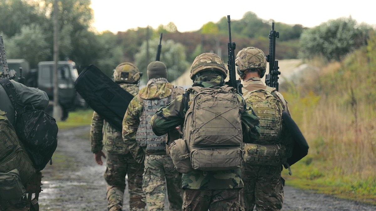 Imaginea prezintă soldați în uniforme de camuflaj, purtând puști și rucsacuri, mergând pe un drum noroios
